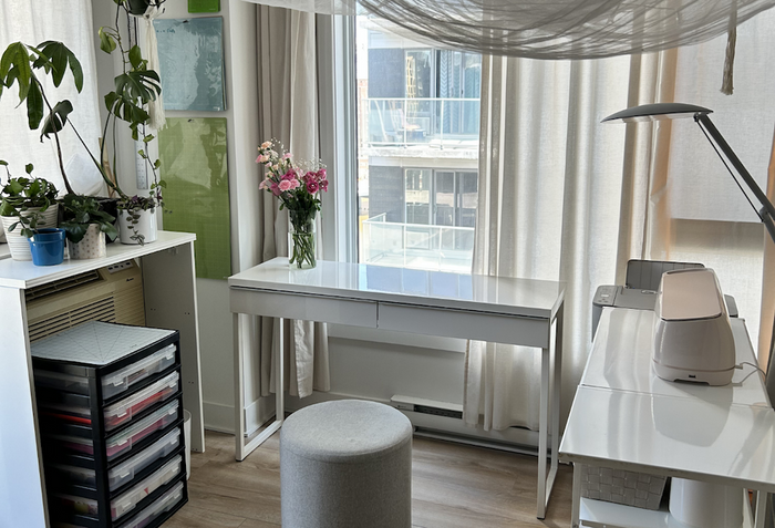 **EN:** Bright and cozy Cricut craft corner set up in a living room. A white desk is placed in front of a large window with sheer curtains, with a Cricut machine on a side table, storage drawers for materials, and a small stool. Plants, hanging fabric, an